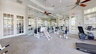 Located at Hawthorne Groves in Orlando, Florida, this fitness center offers a bright and well-appointed space designed to support a full range of workout routines. The room is bathed in natural light, courtesy of large multi-pane windows that line the walls and provide views of the surrounding greenery, creating an open and inviting atmosphere. Ceiling fans are strategically placed throughout, ensuring comfortable airflow during workouts.

The interior features neutral-toned carpet tiles underfoot and crisp white walls accented by tray ceiling details that add architectural interest. Full-length mirrors line one side of the room, visually expanding the space and allowing users to monitor their form during exercise.

The fitness center is comprehensively equipped with both cardio and strength training equipment. Cardio offerings include a treadmill, an elliptical cross-trainer, and a stationary recumbent bike, while the strength training area boasts multiple cable and weight machines suited for a variety of muscle groups. French doors provide direct access to the outdoor area, likely connecting to the broader community amenity spaces. Recessed lighting complements the abundant natural light, ensuring the space remains well-lit at any hour. Overall, the fitness center presents a clean, functional, and welcoming environment for residents.