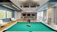 This stylish amenity lounge is thoughtfully designed for recreation and relaxation, offering a dynamic blend of entertainment and comfort across a single level. The space features warm hardwood-style flooring throughout, set beneath a striking two-tone ceiling with teal-painted recessed sections accented by sleek linear LED lighting and recessed spotlights, creating a contemporary and energetic atmosphere.

At the heart of the room, a green felt pool table and a ping-pong table serve as centerpieces, complemented by a foosball table for additional entertainment. A dark tile fireplace with a mounted flat-screen TV above it anchors one end of the room, providing a cozy focal point. Additional wall-mounted TVs are positioned throughout to ensure sightlines from every corner of the space.

A dedicated lounge area lines one wall, featuring white upholstered chairs with gold-accented frames arranged beneath bold green abstract artwork on teal-painted walls. On the opposite end, floor-to-ceiling windows flood the space with natural light and frame views of lush palm trees and a landscaped outdoor area. This windowed nook hosts high-top bistro tables and chairs alongside a decorative gold geometric wall panel, making it an ideal casual gathering spot. The overall design is polished, vibrant, and well-suited for social interaction.