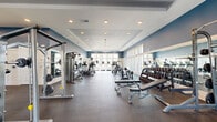 This expansive fitness center offers a generously proportioned, single-floor layout designed to accommodate a full range of workout activities. The interior features a sophisticated two-tone color scheme, with deep navy blue upper walls contrasting against crisp white wainscoting below, creating a clean and energizing atmosphere. A tray ceiling with recessed lighting runs through the main exercise area, complemented by a ceiling fan that enhances air circulation throughout the space. Large, multi-pane windows line the perimeter walls, flooding the facility with abundant natural light and offering views of the surrounding landscape. The durable, speckled rubber flooring provides both safety and comfort underfoot. Equipment is thoughtfully organized throughout the space: a dedicated free weights section features a full dumbbell rack with adjustable benches and wall-mounted mirrors, while a separate strength training zone houses cable machines, a functional training rig with TRX suspension straps, and various selectorized machines. The cardio section includes a row of treadmills, ellipticals, and stair climbers positioned to take advantage of the window views. Wall art adds a finishing touch to the entrance area, contributing to the polished, professional ambiance of this well-appointed fitness facility.