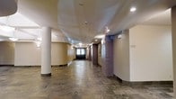 This single-floor space presents a well-appointed interior corridor that combines classic architectural elements with a clean, functional design. The hallway is characterized by its expansive polished stone tile flooring, which features a mix of earthy tones that adds depth and texture underfoot. Smooth, rounded white columns line the passage, offering a refined structural presence, while contrasting accent pillars clad in brick introduce a warm, rustic touch that breaks up the corridor's length with visual interest. The ceiling is finished in white and fitted with a combination of recessed downlights and wall-mounted sconce fixtures, ensuring the space is evenly and warmly illuminated throughout. At the far end of the corridor, a set of dark-framed double doors serves as a focal point, drawing the eye and providing a clear sense of direction. Wayfinding signage is visible along the walls, indicating this is a shared or public-access corridor within a larger building. The overall ambiance is orderly and welcoming, with a neutral color palette of warm beige walls and stone flooring creating a cohesive, professional atmosphere suited to a multi-use or residential building environment.