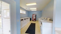 This single-story communal laundry facility offers a clean and functional environment designed to accommodate multiple users simultaneously. The room is painted in a soft, light blue that contributes to an open and airy feel. Rows of white top-load washers and dryers line both sides of the room, providing ample capacity for residents. Overhead, large fluorescent light fixtures illuminate the space evenly, ensuring excellent visibility throughout. A black rubber runner mat runs along the center of the floor, offering a practical and safe walking path between the machines. A Coca-Cola beverage vending machine is positioned at the far end of the room, adding a convenient amenity for users during their laundry visits. A small window near the top of one wall allows a modest amount of natural light to filter in, complementing the bright overhead lighting. The light-toned flooring and white appliances reinforce the facility's clean aesthetic. Overall, the space is straightforward and utilitarian in design, prioritizing functionality and ease of use, making it a practical shared resource for the surrounding residential community.