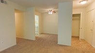 This single-story space features an open floor plan defined by two structural columns that frame the main living area, adding architectural interest while subtly dividing the space. The living area is finished with wall-to-wall carpet in a warm beige tone, contributing to a cozy and comfortable atmosphere throughout. Neutral cream-colored walls keep the space feeling bright and airy, providing a versatile backdrop for a variety of interior styles.

A ceiling fan with an integrated light fixture is centrally positioned in the main living area, offering both functional lighting and air circulation. The structural columns create a natural visual separation between the living space and the adjacent areas, including a glimpse into what appears to be a kitchen and a dedicated laundry nook. The laundry area features a stacked washer and dryer unit, maximizing efficiency within a compact footprint. White cabinetry is visible in the kitchen area, suggesting clean, functional storage. The overall layout is straightforward and practical, making efficient use of the available space on a single level.