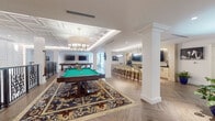 Located at 5400 Broken Sound Blvd NW in Boca Raton, Florida, this elegantly designed single-floor amenity space offers a diverse range of recreational and social environments unified by a refined, club-inspired aesthetic. Herringbone-patterned hardwood flooring flows throughout, complemented by crisp white walls, decorative columns, and coffered ceilings with recessed and pendant lighting that create a warm, upscale atmosphere.

The central game room serves as the heart of the space, featuring a full-size billiards table anchored by a bold floral area rug and illuminated by a large drum chandelier beneath an ornate coffered ceiling. Wrought-iron railings with scrollwork detailing add architectural character to this area. Adjacent to the game room, a dedicated shuffleboard room includes dual wall-mounted flat-screen televisions, providing both recreation and entertainment.

A stylish bar area with white subway tile, a large TV, and a row of upholstered bar stools offers an ideal social gathering spot. A comfortable lounge nook features built-in illuminated shelving and plush barrel-back chairs. The screening room is equipped with a projector, large projection screen, and generous seating including sofas and accent chairs, with natural light filtering through draped windows. Glass-paned doors and open corridors connect each area seamlessly, fostering an inviting, resort-like environment.