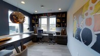 This thoughtfully designed home office delivers a refined and functional workspace on a single floor. The room is anchored by a expansive built-in shelving unit that spans the full width of the wall, offering generous storage for books, decorative objects, and personal mementos. Dark, rich cabinetry and shelving contrast beautifully against the deep navy blue walls, creating a sophisticated and focused atmosphere.

At the heart of the space sits a dual workstation setup, comfortably accommodating two ergonomic office chairs and providing ample desk surface for monitors, printers, and other equipment. A wraparound desk extension along the side wall further maximizes workspace versatility. Recessed ceiling lighting supplements the natural light that pours in through multiple large windows, offering pleasant views of the surrounding landscape and keeping the space bright and energizing throughout the day.

Light-toned, textured carpeting grounds the room and adds warmth underfoot, while a bold piece of wall art introduces a vibrant pop of color and personality. A decorative metallic pendant fixture in the corner adds a sculptural element to the overall aesthetic. The result is a well-appointed office that balances practicality with a polished, contemporary design sensibility.