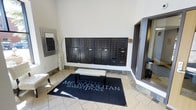 This single-floor space serves as the welcoming lobby and mailroom area for Metropolitan Marketplace. The entrance is defined by neutral-toned tile flooring that lends a clean, polished look to the space. A prominent feature of the lobby is the expansive bank of dark-finished individual mailboxes mounted along the main wall, providing secure mail access for all residents or tenants. A branded Metropolitan Marketplace area rug anchors the center of the room, accompanied by a cushioned bench that offers a convenient spot for sorting mail or brief rest. The walls are finished in a crisp, light tone that enhances the sense of openness and brightness throughout. Decorative elements, including a tall floor vase and a framed mirror, add subtle style to the functional space. A mirrored or glass-paneled entryway is visible to one side, contributing to the airy, open feel of the area. Overall, the lobby strikes a balance between practicality and refined aesthetics, creating a welcoming first impression that reflects the professional character of the Metropolitan Marketplace property.