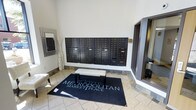 This single-floor space serves as the welcoming lobby and mailroom area for Metropolitan Marketplace. The entrance is defined by neutral-toned tile flooring that lends a clean, polished look to the space. A prominent feature of the lobby is the expansive bank of dark-finished individual mailboxes mounted along the main wall, providing secure mail access for all residents or tenants. A branded Metropolitan Marketplace area rug anchors the center of the room, accompanied by a cushioned bench that offers a convenient spot for sorting mail or brief rest. The walls are finished in a crisp, light tone that enhances the sense of openness and brightness throughout. Decorative elements, including a tall floor vase and a framed mirror, add subtle style to the functional space. A mirrored or glass-paneled entryway is visible to one side, contributing to the airy, open feel of the area. Overall, the lobby strikes a balance between practicality and refined aesthetics, creating a welcoming first impression that reflects the professional character of the Metropolitan Marketplace property.