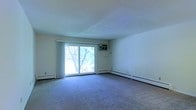 This single-floor space features a generously sized main living area finished with wall-to-wall carpeting in a neutral tone, providing a comfortable and practical foundation. The room is defined by smooth, light-colored walls and a textured ceiling that contributes to a clean, understated aesthetic throughout.

A prominent sliding glass door serves as the focal point of the main living area, allowing an abundance of natural light to fill the space. The door opens to an outdoor area and frames views of mature deciduous trees, creating a natural backdrop that enhances the overall ambiance. White curtain panels flank the door, offering privacy while maintaining the room's bright character.

Climate control is addressed through a wall-mounted air conditioning unit positioned above the sliding door, complemented by baseboard heating units that run along the perimeter of the room — ensuring year-round comfort. The room's layout is open and versatile, offering ample space for a variety of furniture arrangements. Overall, the space presents a functional and straightforward interior with practical features and a connection to the natural surroundings outside.