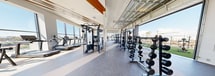 This expansive 2,580 square foot fitness center, located at 3585 Aero Dr in San Diego, offers a comprehensive workout experience. The facility boasts a modern, open-plan design with high ceilings and abundant natural light flooding through floor-to-ceiling windows. The space is thoughtfully divided into distinct zones, each catering to different exercise needs.

The main area features an impressive array of strength training equipment, including multiple squat racks, free weights, and resistance machines. Adjacent to this is a cardio section equipped with the latest treadmills, ellipticals, and stationary bikes. A dedicated functional training area includes suspension trainers, plyometric boxes, and open floor space for bodyweight exercises.

One of the standout features is the indoor-outdoor flow, with large glass doors opening to an artificial turf area. This outdoor space, adorned with palm trees and patio furniture, provides a refreshing environment for al fresco workouts or post-exercise relaxation.

The interior design is sleek and motivating, with a neutral color palette accented by warm wood tones on the ceiling. Strategic lighting, including recessed fixtures and decorative elements, enhances the ambiance. Thoughtful touches like artwork on the walls and conveniently placed storage solutions for personal items contribute to the overall user experience.

This well-equipped, versatile fitness center caters to a wide range of workout preferences, from intensive strength training to cardio sessions and functional fitness, all within a visually appealing and practically designed space.