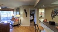 This single-floor residence offers a thoughtfully designed layout that connects living, sleeping, and bathing areas in a cohesive and comfortable flow. The living area is warmly furnished with a cushioned sofa accented by coral throw pillows, a classic floor lamp, and a decorative wall-mounted sculptural figure that adds an artistic touch to the neutral-toned walls. Hardwood flooring runs throughout, lending warmth and continuity to the space. A bar-height counter with upholstered seating provides a casual dining or entertaining option, seamlessly bridging the living area with adjacent spaces. The bedroom, visible through an open doorway, is comfortably appointed with a large bed featuring a dark wood headboard, framed wall art, and soft, layered bedding in neutral tones. Natural light filters in through the windows, contributing to the room's inviting ambiance. The bathroom area, glimpsed through a mirrored vanity reflection, features bright lighting, a well-appointed countertop, and tasteful floral accents. Overall, the space presents a harmonious blend of functional design and warm, welcoming décor suitable for everyday living.