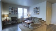 This single-floor space centers around a well-appointed living area designed for comfort and relaxed living. The living room features a generous sectional sofa in a warm taupe fabric, complemented by a coordinating accent chair upholstered in a subtle pattern, creating an inviting and cohesive seating arrangement. A rectangular wooden coffee table anchors the space, providing both function and visual grounding.

The room benefits from natural light streaming through two windows fitted with horizontal blinds, allowing for adjustable brightness throughout the day. The walls are finished in a soft, cool grey tone that lends a calm, contemporary feel to the interior. Dark hardwood-style flooring runs throughout, offering a clean contrast to the lighter furnishings.

Decorative wall art in grid-format mosaic panels adds visual interest on two walls, contributing an artistic, modern touch. A table lamp on a side table provides warm ambient lighting, while a decorative vase with dried branches introduces a natural, organic element. Overall, the space presents a balanced, comfortable atmosphere with thoughtful design details throughout.