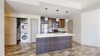 Located on East Sixth Street in Cleveland, Ohio, this single-floor unit offers a well-designed layout that balances open living areas with defined private spaces. The interior is finished with durable stone-look luxury vinyl flooring throughout, lending a cohesive and contemporary feel across every room.

The kitchen serves as a central feature of the unit, anchored by a large island with a white quartz-style countertop, an undermount sink, and a wood-toned base. Dark cabinetry with glass-front upper sections lines the walls, complemented by stainless steel appliances including a refrigerator, range, and microwave. Three decorative pendant lights hang above the island, adding visual interest. Adjacent to the kitchen, a dedicated laundry closet houses a stacked washer and dryer unit behind louvered doors, providing practical convenience.

The living area is generously proportioned, with multiple large windows offering direct views of surrounding downtown Cleveland buildings, filling the space with natural light. Additional rooms, suitable for bedrooms or flex use, each feature windows with urban views and are finished in neutral tones.

The bathroom includes a glass-enclosed walk-in shower with white subway tile, a dark-toned vanity with a granite countertop, and large-format tile flooring. The overall design maintains a clean, modern aesthetic throughout.