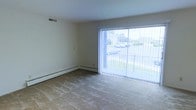 This single-floor residential space offers a clean, functional layout with a comfortable and understated aesthetic. The living area is a standout feature, highlighted by expansive sliding glass doors dressed with vertical blinds that flood the room with natural light and provide an open view of the surrounding greenery and outdoor surroundings. The neutral-toned walls and light-colored carpeted flooring create a calm, welcoming atmosphere that is easy to personalize. Baseboard heating runs along the perimeter of the room, ensuring efficient and consistent warmth throughout the colder months. A ceiling air vent further supports year-round climate control, adding to the practicality of the space. The room's generous proportions allow for a variety of furniture arrangements, making it well-suited for both relaxing and entertaining. The overall design is straightforward and functional, with smooth white ceilings and minimal architectural detailing that lend the space a clean, uncluttered feel. The abundant natural light combined with the outdoor views gives the interior an airy quality that enhances the sense of openness throughout this single-level residence.