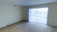 This single-floor residential space offers a clean, functional layout with a comfortable and understated aesthetic. The living area is a standout feature, highlighted by expansive sliding glass doors dressed with vertical blinds that flood the room with natural light and provide an open view of the surrounding greenery and outdoor surroundings. The neutral-toned walls and light-colored carpeted flooring create a calm, welcoming atmosphere that is easy to personalize. Baseboard heating runs along the perimeter of the room, ensuring efficient and consistent warmth throughout the colder months. A ceiling air vent further supports year-round climate control, adding to the practicality of the space. The room's generous proportions allow for a variety of furniture arrangements, making it well-suited for both relaxing and entertaining. The overall design is straightforward and functional, with smooth white ceilings and minimal architectural detailing that lend the space a clean, uncluttered feel. The abundant natural light combined with the outdoor views gives the interior an airy quality that enhances the sense of openness throughout this single-level residence.