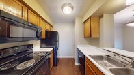 This single-story unit is designed with a functional, straightforward layout that connects several distinct living areas. The galley-style kitchen is efficiently organized, featuring rich oak cabinetry with ample upper and lower storage, light speckled countertops, and a full suite of black appliances including an electric range, over-the-range microwave, refrigerator, and dishwasher. Dark hardwood-style flooring runs throughout the kitchen and extends into the main living areas, adding visual continuity. The living room is notably open, receiving natural light through a sliding glass door that provides direct outdoor access. Adjacent to the living space, a separate dining area is defined by a pendant light fixture overhead. The bedrooms feature wall-to-wall carpeting, ceiling fans, and neutral-toned walls, creating a comfortable, quiet atmosphere. One bedroom includes a window with outdoor views. The bathroom is appointed with an oak vanity, speckled countertop, a four-bulb vanity light bar, a large mirror, and a shower-tub combination. Throughout the unit, textured ceilings and clean white trim provide a cohesive, understated finish.