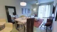 This single-floor space features an open-concept layout that seamlessly connects the dining and living areas, creating a cohesive and functional environment. The dining area is anchored by a striking stone-top table with a marbled finish, paired with dark wood chairs that add warmth and contrast. A modern drum pendant light hangs above, providing focused illumination over the dining surface.

The living area flows naturally from the dining space, centered around a neutral-toned sofa and ottoman arrangement set atop a rich, textured area rug in warm earth tones. A ceiling fan with a brushed metal finish is mounted overhead, contributing to the room's practical comfort. A wire-frame accent chair adds a sculptural touch near the window.

Natural light is a defining feature of the space, streaming in through a large sliding glass door that offers views of the outdoors and creates an airy, open ambiance. Soft blue-gray walls complement the light tones of the furnishings throughout. A curated gallery wall of framed artwork adds visual interest, while a tall floor lamp provides warm supplemental lighting. The overall aesthetic is contemporary, clean, and inviting.