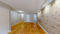 This single-floor unit offers a distinctive blend of industrial character and residential comfort. The main living area is the standout space, featuring a full wall of exposed brick illuminated by track lighting, which adds warmth and texture to the room. Hardwood floors in a rich honey tone run throughout, complementing the neutral gray walls and white trim. A closet and a window providing natural light are also present in this open, versatile space.

The layout extends into a separate kitchen area, equipped with gray cabinetry, a double stainless steel sink, and butcher block-style countertops. A window fitted with horizontal blinds allows natural light into the cooking space. The textured ceiling continues throughout, giving the unit a consistent aesthetic.

The bathroom is cleanly designed with white square tile lining the tub and shower surround, mosaic tile flooring, a wall-mounted sink, a mirrored medicine cabinet, and a lighted vanity fixture above. The overall ambiance of the unit balances raw architectural details—most notably the exposed brick—with practical, well-maintained finishes, making it a functional and visually engaging space.