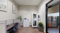 Located at 73 Brownstone Way in Englewood, NJ, this business center offers a thoughtfully designed, single-floor workspace that balances collaborative and focused work environments. The space features warm hardwood-style flooring throughout, neutral-toned walls, and recessed lighting, creating a professional yet welcoming atmosphere.

At the heart of the space is a conference area furnished with a sleek white rectangular table surrounded by modern black mesh chairs, complemented by a large whiteboard mounted on the wall — ideal for meetings and presentations. Black-framed glass doors and interior windows allow natural light to flow through while maintaining visual connectivity between areas.

A dedicated workstation zone features a row of private cubicles equipped with ergonomic chairs and individual desks, providing focused work areas for individual productivity. Botanical-inspired framed artwork adorns the walls, lending a calm, refined aesthetic to the workspace.

A standout feature is the freestanding privacy pod — a self-contained phone booth designed for confidential calls or quiet focus sessions. Nearby, a print station with a multifunction printer and storage cabinet adds practical utility. Lush potted greenery is placed throughout, enhancing the overall ambiance. This business center is a comprehensive, fully equipped workspace suited for a variety of professional needs.