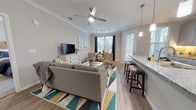 Located in Charleston, SC, this single-floor residence at Ashley Gardens offers a thoughtfully designed open-concept layout with modern finishes throughout. The kitchen serves as a central hub, featuring gray shaker-style cabinetry, granite countertops, stainless steel appliances, and a large island with pendant lighting and bar seating — ideal for both cooking and entertaining. Wood-look flooring flows seamlessly from the kitchen into the living area, which is brightened by multiple windows dressed in navy curtains, a ceiling fan with track lighting, and crown molding that adds an elegant touch. The living space opens to a private balcony, where a wrought iron railing frames tranquil views of a pond and lush greenery. The bedrooms offer plush carpeting, ceiling fans, ample closet space, and generous natural light. Both bathrooms are finished with granite-topped gray vanities and large mirrors; one features a shower/tub combo. The neutral gray and white color palette, crown molding, and consistent finishes create a cohesive and polished ambiance throughout this well-designed Charleston residence.