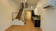 This two-story space showcases a thoughtfully designed open-plan layout that combines modern aesthetics with functional living. The main level features warm bamboo-style hardwood flooring that runs throughout, creating a cohesive and inviting foundation for the space.

The kitchen is positioned along one wall and is outfitted with dark cabinetry, granite or stone countertops, and stainless steel appliances including a refrigerator and microwave. A dark tile backsplash adds texture and contrast against the white walls, while recessed and pendant lighting illuminate the workspace effectively.

A standout architectural feature is the open-riser staircase with metal railings, which connects both floors while contributing an industrial edge to the overall design. The staircase is positioned to allow sightlines through to the upper level, enhancing the sense of vertical space and openness. The high ceiling height amplifies this effect, making the main level feel expansive.

The white walls throughout provide a clean, neutral backdrop that complements the darker finishes. A hallway visible toward the rear of the main level leads to additional rooms, including what appears to be a bathroom, offering a practical and well-organized floor plan.