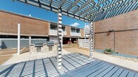This communal outdoor space is situated within a multi-story brick building complex, designed around a central courtyard that encourages gathering and outdoor living. The focal point of the space is a generously sized wood-plank deck finished in a cool gray tone, anchored by a striking white geometric pergola overhead that casts dramatic shadow patterns across the surface and provides partial shade throughout the day.

Adjacent to the deck is a covered lounge pavilion with a vaulted ceiling, ceiling fan, and large open-air openings on multiple sides, furnished with blue cushioned seating and metal accent tables, creating a comfortable, shaded retreat. The pavilion offers direct sightlines to a sculptural spiral staircase and the surrounding brick architecture, reinforcing the cohesive mid-century aesthetic of the complex.

A built-in outdoor kitchen station with a gas grill sits atop a stacked stone base nearby, offering a convenient amenity for residents. The surrounding courtyard features low-maintenance gravel landscaping with native plantings and stone accents, bordered by white horizontal-slatted privacy screens. Covered parking is accessible from the courtyard, and the overall layout benefits from abundant natural light and an open-sky exposure that enhances the sense of openness and connection to the outdoors.