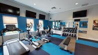 This fitness center is designed to support a comprehensive workout experience within a single, open-plan room. The space is defined by bold cobalt-blue accent walls contrasted against light gray upper walls and white crown molding, creating an energetic yet clean aesthetic. Recessed lighting throughout ensures the room is well illuminated, complemented by natural light from multiple windows along the perimeter.

The cardio zone features a row of treadmills and elliptical machines positioned along the windowed wall, each equipped with individual screens and accompanied by wall-mounted flat-screen televisions overhead for entertainment during workouts. A rowing machine is also available for low-impact cardiovascular training.

The strength training area includes a fully stocked dumbbell rack with a range of weights, a flat utility bench, and a multi-function cable resistance machine suitable for a variety of exercises. A large wall mirror spans this section, enhancing the sense of space and allowing users to monitor their form.

A dedicated functional training corner houses an assortment of stability balls, medicine balls, and resistance accessories stored on a upright rack. Motivational fitness artwork is displayed on the walls. The flooring consists of multi-toned rubber tiles in blue, gray, and tan, offering both durability and traction. The space also includes a restroom accessible directly from the fitness area.