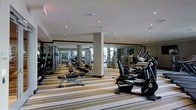 This fitness center offers a comprehensive workout environment designed to accommodate a variety of exercise routines. The space is generously proportioned, with recessed pot lights embedded in a clean white ceiling that provide bright, even illumination throughout. Large floor-to-ceiling mirrors line the walls, visually expanding the room and allowing users to monitor their form during exercise. Structural columns are integrated into the layout without disrupting the flow of the space.

The flooring features a distinctive striped carpet in neutral tones, adding a refined aesthetic while providing a degree of cushioning underfoot. The equipment selection is extensive, including upright and recumbent stationary bikes, elliptical trainers, and a variety of strength and resistance machines arranged in organized rows. Free weight benches and adjustable equipment further enhance the versatility of the facility.

The overall ambiance is clean, modern, and functional, with a layout that ensures ample space between machines for comfortable movement. The mirrored walls also reflect natural light from nearby windows, contributing to a bright and energizing atmosphere well-suited for fitness activities.