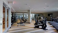 This fitness center offers a comprehensive workout environment designed to accommodate a variety of exercise routines. The space is generously proportioned, with recessed pot lights embedded in a clean white ceiling that provide bright, even illumination throughout. Large floor-to-ceiling mirrors line the walls, visually expanding the room and allowing users to monitor their form during exercise. Structural columns are integrated into the layout without disrupting the flow of the space.

The flooring features a distinctive striped carpet in neutral tones, adding a refined aesthetic while providing a degree of cushioning underfoot. The equipment selection is extensive, including upright and recumbent stationary bikes, elliptical trainers, and a variety of strength and resistance machines arranged in organized rows. Free weight benches and adjustable equipment further enhance the versatility of the facility.

The overall ambiance is clean, modern, and functional, with a layout that ensures ample space between machines for comfortable movement. The mirrored walls also reflect natural light from nearby windows, contributing to a bright and energizing atmosphere well-suited for fitness activities.