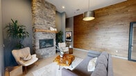 Located in Maple Valley, Washington, this well-appointed single-floor space offers a thoughtfully designed layout that blends modern aesthetics with functional versatility. The living area immediately draws attention with its dramatic floor-to-ceiling stacked stone fireplace, anchored by a raw wood mantel and flanked by a warm wood-paneled accent wall. Soaring ceilings with exposed dark wood beams add architectural character, while large floor-to-ceiling windows and glass entry doors invite abundant natural light and views of the surrounding exterior.

The living space accommodates multiple seating arrangements, featuring plush sectional sofas, accent chairs, and organic live-edge coffee tables atop area rugs over large-format tile flooring. A wall-mounted flat-screen display is integrated into the wood accent wall, enhancing the room's media capabilities.

The kitchen is sleek and fully equipped, showcasing warm wood cabinetry, gray quartz countertops, a textured tile backsplash, and stainless steel appliances including a refrigerator, microwave, and dishwasher. A spacious island with bar seating serves as a central gathering point, illuminated by globe pendant lighting.

Flexible glass-paneled folding and swing doors lead to additional private rooms, offering adaptable spaces suited for meetings or focused work, completing this versatile and stylish environment.