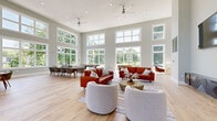Located at 805 Quince Orchard Blvd in Gaithersburg, MD, this single-story community clubhouse offers a generously sized, open-plan interior designed for both relaxation and social gatherings. The space is defined by its dramatically tall ceilings and an expansive wall of oversized, white-framed windows that flood the interior with natural light and frame views of the surrounding greenery and neighborhood.

Light hardwood flooring runs throughout, complementing the soft sage-gray wall tones and crisp white trim to create a clean, contemporary aesthetic. The main area is thoughtfully arranged into distinct functional zones. A comfortable lounge area features modern leather seating and accent chairs gathered around a central coffee table, while a spacious dining area accommodates a large table with ample seating for group events. A recreation zone includes game tables, adding an entertaining element to the space.

A service counter with a pass-through window provides additional functionality, suggesting club or management services on-site. Double glass entry doors with secure access hardware offer a welcoming yet controlled entrance, with views extending to an outdoor landing and steps. Recessed lighting and ceiling fans complement the natural light, ensuring the space remains comfortable year-round. Overall, the clubhouse exudes a bright, airy, and inviting ambiance well-suited for community use.
