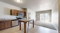 Located at 218 19th St SE in Watertown, South Dakota, this 2-bedroom, 2-bathroom unit offers a practical and well-appointed single-floor layout. The open-concept kitchen and living area serves as the heart of the unit, where light vinyl plank flooring in the kitchen transitions seamlessly into carpeted living space. The kitchen is equipped with a full suite of stainless steel appliances — including a refrigerator, electric range, built-in microwave, and dishwasher — complemented by warm medium-tone cabinetry and granite-style countertops. A breakfast bar island adds additional prep and dining space. The living area benefits from a sliding glass door that opens to a private balcony, allowing for natural light and outdoor access. Both bedrooms are carpeted and feature neutral tones; the primary bedroom is generously sized and connects to a bathroom with a walk-in closet and a vanity with matching cabinetry. The second bathroom includes a full tub-shower combination. A dedicated laundry room houses a full-size washer and dryer. Textured ceilings and baseboard heating are consistent throughout, adding to the unit's functional, comfortable character.