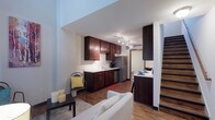 This two-story space offers a thoughtfully designed open-concept layout that seamlessly connects the living and kitchen areas on the main floor. The kitchen is well-appointed with rich dark-wood cabinetry, light countertops, and stainless steel appliances, including a refrigerator and dishwasher. Track lighting runs along the ceiling, providing focused illumination throughout the cooking space. A breakfast bar-style counter separates the kitchen from the adjacent living area, creating a natural transition between the two zones. The living area is bright and welcoming, featuring wood-look flooring that extends throughout the main level, adding warmth and continuity to the space. A light-toned sofa and area rug anchor the living zone, while tall ceilings contribute to an open, airy feel. A staircase with dark wood treads and a clean, modern design rises along one wall, leading to the upper floor. The overall aesthetic is contemporary, combining dark wood accents with neutral whites and warm flooring tones to create a balanced and functional living environment.