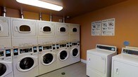 This single-floor shared laundry facility is designed with practicality and efficiency in mind. The room features warm amber-painted walls that create a welcoming atmosphere, complemented by light wood-style flooring and bright overhead fluorescent lighting that ensures excellent visibility throughout the space.

The room is outfitted with a row of stacked front-loading washer and dryer units, providing ample laundry capacity for multiple users. Each stacked unit pairs a front-loading washing machine on the bottom with a corresponding dryer on top, maximizing the use of vertical space. In addition to the stacked units, a standalone top-loading washing machine is positioned along the adjacent wall, offering users a variety of machine options.

Instructional guides are clearly posted on the wall, including easy-to-follow washing and operational guides, making the facility accessible and user-friendly for all. Exposed ventilation ductwork runs along the upper wall behind the machines, reflecting the room's utilitarian design. A floor drain is centrally located, adding a practical and hygienic element to the space.

Overall, this laundry facility is thoughtfully organized to accommodate multiple users simultaneously, balancing functionality with a clean, approachable aesthetic.