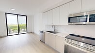 Located at 1155 NE 126th St in North Miami, FL, this single-floor unit offers a clean, modern aesthetic throughout its thoughtfully designed layout. The open-plan living and kitchen area is anchored by light wood flooring that runs seamlessly across the space, creating a bright and cohesive feel. The contemporary kitchen features flat-front cabinetry in white and warm greige tones extending from floor to ceiling, providing ample storage. A full suite of stainless steel appliances — including an electric range, built-in microwave, dishwasher, and refrigerator — is paired with a sleek single-basin sink and white quartz countertops. The living area benefits from a large floor-to-ceiling window that frames outdoor views of the surrounding North Miami neighborhood, filling the room with natural light. A dedicated bedroom area also features an expansive floor-to-ceiling glazed door, further enhancing the connection to the outdoors and maximizing daylight. The bathroom is accessed via a carpeted hallway and showcases warm wood-toned cabinetry, a wide backlit mirror, and a window that introduces natural light. The overall design is characterized by a minimalist palette of white walls, neutral tones, and clean lines, resulting in a contemporary and functional living environment.