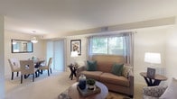 This single-floor residence offers a thoughtfully designed layout that balances comfort and functionality across its key living spaces. The living room serves as the heart of the home, featuring plush carpeting, large windows dressed with sheer curtains and blinds, and a sliding glass door that invites natural light and outdoor views. The open arrangement easily accommodates both a seating and a workspace area, making the room highly versatile. Adjacent to the living room, the dining area benefits from the same abundant natural light through a full-length vertical blind slider, and is anchored by a pendant light overhead, creating an inviting atmosphere for everyday meals. The kitchen is equipped with white cabinetry, granite-style countertops, a stainless steel sink, and dark hardwood-style flooring, providing a practical and attractive space for cooking. The residence includes two well-sized bedrooms, each featuring soft carpeting, generous closet storage, and windows that allow natural light to filter in. The bathroom is finished with classic white tile wainscoting, a wide vanity with a large mirror, and clean, bright fixtures throughout. Overall, the space presents a warm, neutral palette and a comfortable, livable ambiance.