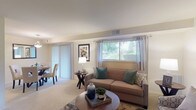 This single-floor residence offers a thoughtfully designed layout that balances comfort and functionality across its key living spaces. The living room serves as the heart of the home, featuring plush carpeting, large windows dressed with sheer curtains and blinds, and a sliding glass door that invites natural light and outdoor views. The open arrangement easily accommodates both a seating and a workspace area, making the room highly versatile. Adjacent to the living room, the dining area benefits from the same abundant natural light through a full-length vertical blind slider, and is anchored by a pendant light overhead, creating an inviting atmosphere for everyday meals. The kitchen is equipped with white cabinetry, granite-style countertops, a stainless steel sink, and dark hardwood-style flooring, providing a practical and attractive space for cooking. The residence includes two well-sized bedrooms, each featuring soft carpeting, generous closet storage, and windows that allow natural light to filter in. The bathroom is finished with classic white tile wainscoting, a wide vanity with a large mirror, and clean, bright fixtures throughout. Overall, the space presents a warm, neutral palette and a comfortable, livable ambiance.