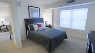This single-floor space showcases a thoughtfully designed bedroom that balances comfort and style. The room is anchored by a striking dark-paneled headboard paired with a queen-sized bed dressed in a charcoal quilted comforter and an assortment of coordinating pillows, including accents in rust and gray tones. A nightstand with a softly glowing table lamp sits beside the bed, adding warmth to the space.

The room features neutral wall tones and light-colored carpeting that contribute to a calm, cohesive atmosphere. A large window fitted with horizontal blinds allows natural light to fill the space, enhancing its open and airy feel. The window's placement provides a bright focal point along one wall, complementing the room's overall layout.

Crown molding details along the ceiling add a subtle architectural refinement, while a framed piece of wall art above the headboard introduces a decorative element. A ceiling vent is visible, indicating a central air system. The room's layout offers ample space around the bed, making it functional and comfortable for everyday use.