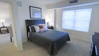 This single-floor space showcases a thoughtfully designed bedroom that balances comfort and style. The room is anchored by a striking dark-paneled headboard paired with a queen-sized bed dressed in a charcoal quilted comforter and an assortment of coordinating pillows, including accents in rust and gray tones. A nightstand with a softly glowing table lamp sits beside the bed, adding warmth to the space.

The room features neutral wall tones and light-colored carpeting that contribute to a calm, cohesive atmosphere. A large window fitted with horizontal blinds allows natural light to fill the space, enhancing its open and airy feel. The window's placement provides a bright focal point along one wall, complementing the room's overall layout.

Crown molding details along the ceiling add a subtle architectural refinement, while a framed piece of wall art above the headboard introduces a decorative element. A ceiling vent is visible, indicating a central air system. The room's layout offers ample space around the bed, making it functional and comfortable for everyday use.