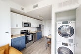 Located at 50-54 Sussex Avenue in Newark, NJ, this thoughtfully designed one-bedroom, one-bathroom residence offers a well-utilized layout with modern finishes throughout. The open kitchen flows seamlessly into the living area, featuring two-tone cabinetry with white upper cabinets and navy blue lower cabinets, granite countertops, a white subway tile backsplash, and a full suite of stainless steel appliances. Light wood-style flooring runs throughout the main living areas, contributing to a bright and cohesive aesthetic. A dedicated dining nook and in-unit stacked washer and dryer add everyday convenience. The spacious bedroom is a standout feature, showcasing a dramatic curved wall of floor-to-ceiling black-framed windows that bathe the room in natural light and frame sweeping views of the Newark skyline and surrounding cityscape. A private balcony is accessible directly from the bedroom. Two open closets provide generous storage. The bathroom is finished with large-format dark slate tile flooring, white cabinetry, and a granite-topped vanity. The overall ambiance is clean, contemporary, and light-filled, making efficient use of every square foot in this modern urban residence.