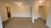 This single-story space presents a straightforward, functional layout with an open design that connects the main living area to adjacent spaces. The living room is finished with wall-to-wall neutral-toned carpet, offering a warm and comfortable underfoot feel throughout. A ceiling fan with an integrated light fixture serves as the central overhead lighting source, suspended from a textured ceiling that adds subtle visual character to the room.

The walls are painted in a clean, neutral white, providing a blank canvas adaptable to various interior styles. A structural column or partial wall divides the main living area from a secondary space, creating a natural sense of separation while maintaining an open feel. A louvered closet door is visible along the back wall, offering practical storage within the living area.

The layout flows into an adjoining area, partially visible to the right, suggesting additional living or utility space. Multiple electrical outlets are positioned along the walls at standard heights, ensuring convenient access throughout. The overall ambiance is clean, neutral, and move-in ready, suited for a range of furnishing arrangements.