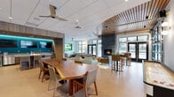 Located at 435 Alexander Loop in Eugene, Oregon, this thoughtfully designed single-floor community lounge offers a generously proportioned open-plan gathering space that seamlessly blends comfort and contemporary style. The interior is anchored by a striking live-edge wood communal table surrounded by modern dining chairs, serving as an ideal centerpiece for social interaction. A curved wood-slat ceiling detail adds architectural warmth and visual interest, complementing the light herringbone-patterned hardwood flooring throughout.

Multiple distinct seating zones encourage both casual lounging and more intimate conversation. A plush sofa arrangement is anchored by a patterned area rug, while teal accent chairs and earth-toned sofas provide color and comfort. A floor-to-ceiling stone fireplace serves as a dramatic focal point, creating a cozy atmosphere in the lounge area. High-top bar seating near the windows offers an additional social spot with views to the outdoor courtyard.

Large black-framed windows and glass garage-style doors flood the space with natural light and provide direct access to the outdoor patio area. A built-in kitchenette nook, enclosed by warm wood-paneled walls with teal accent lighting, features a bar-height island and mounted screens for entertainment. A large-format display television is mounted prominently for communal viewing, completing this versatile and well-appointed amenity space.