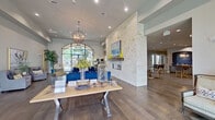 This sophisticated single-floor community space combines modern design with warm, inviting details throughout its open layout. The expansive main lounge area immediately impresses with soaring ceilings, rich hardwood flooring, and dramatic floor-to-ceiling arched windows framed in sleek black steel, flooding the interior with natural light and offering views to the outdoors. A striking floor-to-ceiling stacked stone fireplace serves as the central architectural focal point, topped with a wood mantle and framed artwork, anchoring the living area where plush cobalt blue sofas and layered rugs create an inviting gathering space. Pendant globe chandeliers with an industrial-chic aesthetic illuminate both the lounge and reception areas. A well-appointed kitchen features slate-blue shaker cabinetry, dark granite countertops, stainless steel appliances including a range with a chimney hood, and a large window overlooking the exterior. A dining area connects seamlessly to a distinctive beverage station set within a stone archway, accented by vibrant cobalt mosaic tile and a decorative chandelier. Additional seating areas with coordinating grey armchairs and curated artwork complete the refined, coastal-inspired aesthetic of this thoughtfully designed communal space.