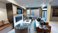 This single-floor space centers around a well-appointed living area that blends contemporary design with comfortable, eclectic furnishings. The focal point of the room is a sleek, linear electric fireplace framed by a white tile surround and dark wood accents, above which a large wall-mounted flat-screen television is installed. Recessed lighting in the ceiling complements the natural light that floods the space through multiple large windows, offering views of the surrounding urban environment and an adjacent outdoor area.

The seating arrangement is both stylish and inviting, featuring a cognac leather sofa accented with deep teal throw pillows, a tufted grey velvet accent chair, and a tufted dark leather chair. A rectangular coffee table with a light wood top and dark metal frame anchors the seating area, which is grounded by a multicolored, abstract area rug that introduces bold pops of color against the neutral grey walls and flooring. A small side table adds functional surface space near the seating. The overall ambiance is modern and curated, combining warm textures with cool tones for a sophisticated yet welcoming atmosphere.