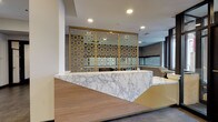 This thoughtfully designed amenity floor makes a strong first impression with a grand lobby entrance featuring large-format stone tile flooring, textured accent walls, and a striking marble-fronted reception desk adorned with an ornate gold geometric screen divider. The open lobby leads to a well-appointed co-working area with workstations positioned near windows that invite natural light. An elevator and flanking stairways with stone treads provide seamless vertical access throughout the building. The lounge area is a vibrant social hub, furnished with a mix of teal accent chairs, a bold patterned ottoman, and a long upholstered banquette in warm yellow tones, complemented by stylish wall-mounted sconce lighting. Adjacent to the lounge, a cozy TV room features a built-in wall-mounted display, a full bookshelf library, plush seating, and a marble-top coffee table, creating an inviting retreat. The game room offers a centerpiece billiards table in olive green, bar-height seating along a built-in counter, and access to a separate lounge space with yellow seating visible beyond glass partitions. A private conference room rounds out the space, outfitted with a solid wood table, teal upholstered chairs, and a dramatic circular pendant light fixture. Throughout, the design blends warm wood tones, marble accents, and bold pops of color for a polished, contemporary ambiance.