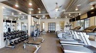This fitness center offers a thoughtfully designed workout environment that balances functionality with refined aesthetics. The single-floor layout provides an open, spacious feel that accommodates a wide range of exercise equipment without feeling crowded. The ceiling is a standout architectural feature, showcasing coffered tray detailing with rich wood paneling and recessed lighting, complemented by elegant pendant-style ceiling fans that maintain comfortable airflow throughout the space. Full-length mirrors line the walls, amplifying the sense of openness and allowing users to monitor their form during workouts. The flooring consists of durable, cushioned exercise matting suited for high-traffic fitness use. Along one side of the room, a row of cardio machines — including ellipticals and treadmills — is neatly arranged, while the opposite side hosts a comprehensive selection of strength training equipment, including cable machines and a full dumbbell rack. Built-in storage cubbies near the entrance provide convenient organization for personal items. Natural light filters in through windows, brightening the interior and creating an inviting atmosphere. Overall, the space strikes a balance between upscale design and practical, performance-focused amenities.