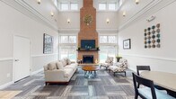 This single-floor communal space is defined by its dramatically tall ceilings and open, airy layout that immediately conveys a sense of grandeur. The main lounge area serves as the heart of the space, anchored by a striking full-height brick fireplace and chimney that extends to the ceiling, with a mounted flat-screen television above the mantle. Multiple tiers of windows — including clerestory windows near the roofline — flood the room with abundant natural light and offer views of the surrounding outdoor area, including a glimpse of what appears to be a pool beyond the glass doors.

The lounge is furnished with a neutral-toned sofa, patterned accent chairs, and a round wood coffee table, all arranged on a multi-toned modular carpet that adds texture and visual interest. Crown molding and wainscoting along the lower walls lend a refined, classic architectural character throughout.

A secondary seating and dining area near the entrance features a small round table with chairs beneath an arched ceiling detail, offering an intimate spot for casual meetings. The connected kitchenette is equipped with warm wood cabinetry, a marble-style countertop, stainless steel sink, refrigerator, microwave, and coffee maker — providing convenient refreshment amenities for occupants.