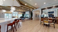 Located at 7035 Broad Way in Davenport, Florida, this single-floor community clubhouse offers a thoughtfully designed collection of amenity spaces that blend modern aesthetics with practical functionality. The expansive main lounge serves as the heart of the facility, featuring warm wood-look flooring, exposed decorative ceiling beams, recessed lighting, and a mix of comfortable seating arrangements. A grand piano adds a sophisticated touch, while French doors open directly to an outdoor patio, filling the space with natural light and views of the surrounding community. A long, white quartz bar island with upholstered barstools anchors the social hub, complemented by globe pendant lighting and a coffee and beverage station set within rich wood cabinetry. A bright dining area offers communal seating alongside floor-to-ceiling windows that frame lush outdoor views, enhanced by a striking textured accent wall. The game room provides a bold contrast with dark shiplap walls, a full-size pool table, wall-mounted television, and patterned flooring. The fitness center is well-equipped with strength machines, stationary bikes, and cardio equipment, energized by a vibrant motivational mural and tall windows that bring in natural light. Throughout, the design balances contemporary style with inviting warmth, creating a versatile space suited for both relaxation and activity.