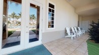 This single-story property offers a quintessential Florida lifestyle, seamlessly blending indoor comfort with stunning outdoor living. The covered patio serves as a welcoming entryway, finished with light tile flooring and framed by French-style glass doors and multi-pane windows that fill the interior with natural light. A pair of classic white Adirondack chairs invites relaxation just outside the entrance, offering a shaded spot to enjoy the warm surroundings.

Stepping outside, the grounds are immaculately landscaped with lush green turf, vibrant tropical plantings, decorative ceramic urns, and native Florida flora that create a resort-like atmosphere. Tall palm trees line the property, providing shade and that signature tropical character. A paver walkway leads directly to a sparkling community pool, which is surrounded by a broad tiled deck and additional tropical greenery, making it an ideal space for outdoor recreation and leisure.

The architectural style reflects classic Florida Mediterranean influences, with stucco exterior walls, terracotta-toned rooflines visible in the background, and clean white trim detailing. The combination of thoughtfully designed outdoor spaces, abundant natural light, and lush poolside setting makes this property a truly inviting and functional retreat suited for the Florida lifestyle.