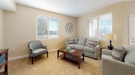 This single-floor residence offers a thoughtfully designed layout with warm, neutral tones and an abundance of natural light throughout. The living room serves as a welcoming centerpiece, featuring plush carpeting, multiple windows with adjustable blinds that frame outdoor views, and a comfortable seating arrangement centered around a wood coffee table. A decorative wall clock adds a charming accent to the space.

The kitchen is designed for functionality, featuring rich cherry wood cabinetry, granite-style countertops, a double stainless steel sink, and white appliances including a refrigerator, range, and dishwasher. Wood-look flooring runs through the kitchen and flows seamlessly into the adjacent dining nook, where a round wooden table with chairs sits beside a bright window — ideal for everyday meals.

The primary bedroom offers a serene retreat with soft carpeting, a walk-in closet, and a built-in desk area with overhead cabinetry, providing practical workspace. A second bedroom, also carpeted and filled with natural light, offers comfortable accommodations with ample room for furnishings.

The bathroom is finished with a rich wood vanity, granite-style countertop, a large mirror, and dark wood-look flooring, complemented by a shower-tub combination. Throughout the residence, the consistent use of warm beige walls and quality finishes creates a cohesive and inviting atmosphere.