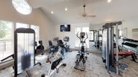 This fitness center is designed to support a comprehensive workout experience within a single, open-plan space. The room's most striking architectural feature is its vaulted ceiling, which rises to an impressive peak and is accentuated by a circular skylight that floods the interior with natural light. Recessed lighting and a ceiling fan provide additional comfort and illumination throughout the space.

The flooring features a neutral, patterned carpet tile that extends across the entire area, providing a comfortable surface for exercise. Large windows along the walls invite views of the surrounding greenery and allow abundant daylight to enhance the open, airy atmosphere.

The fitness center is thoughtfully equipped with a wide range of exercise equipment. Cardio options include treadmills, stationary bikes, a recumbent bike, and an elliptical trainer, while a multi-station cable machine and a rack of dumbbells cater to strength training needs. A wall-mounted flat-screen television, a body weight scale, and wall mirrors add functionality and convenience.

A set of double glass doors opens directly to an outdoor deck, seamlessly connecting the interior to the exterior environment. Built-in storage cabinetry and a hand sanitizer dispenser round out the practical amenities, making this a fully functional and inviting fitness facility.