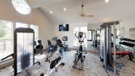 This fitness center is designed to support a comprehensive workout experience within a single, open-plan space. The room's most striking architectural feature is its vaulted ceiling, which rises to an impressive peak and is accentuated by a circular skylight that floods the interior with natural light. Recessed lighting and a ceiling fan provide additional comfort and illumination throughout the space.

The flooring features a neutral, patterned carpet tile that extends across the entire area, providing a comfortable surface for exercise. Large windows along the walls invite views of the surrounding greenery and allow abundant daylight to enhance the open, airy atmosphere.

The fitness center is thoughtfully equipped with a wide range of exercise equipment. Cardio options include treadmills, stationary bikes, a recumbent bike, and an elliptical trainer, while a multi-station cable machine and a rack of dumbbells cater to strength training needs. A wall-mounted flat-screen television, a body weight scale, and wall mirrors add functionality and convenience.

A set of double glass doors opens directly to an outdoor deck, seamlessly connecting the interior to the exterior environment. Built-in storage cabinetry and a hand sanitizer dispenser round out the practical amenities, making this a fully functional and inviting fitness facility.